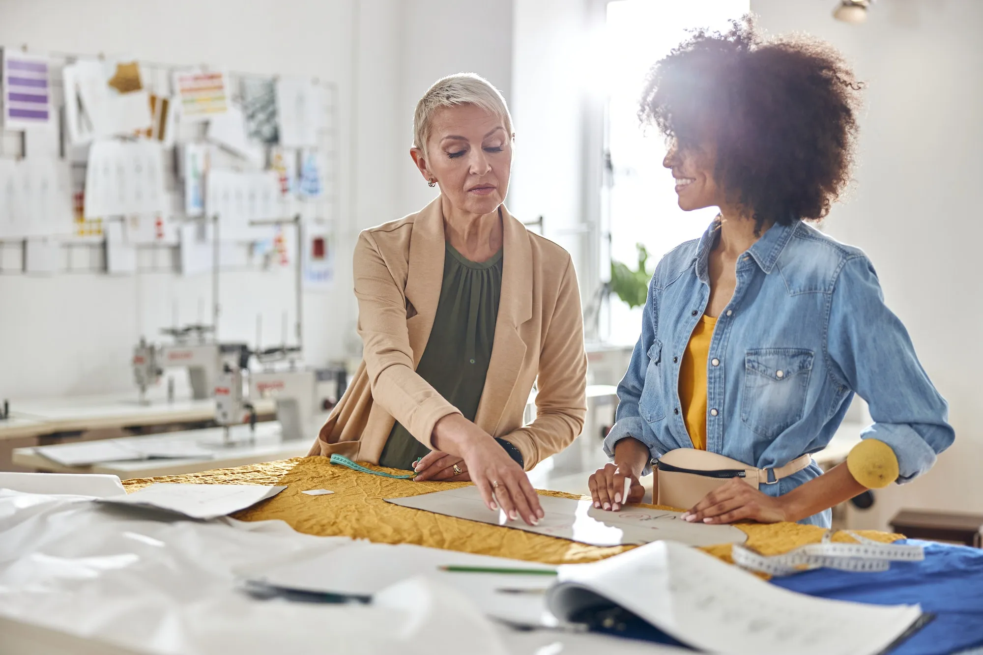 Mature lady boss checks sewing patterns made by young African-American seamstress at cutting table
